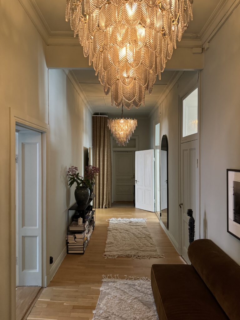 Long hallway furnished in an elegant way with chandeliers, several rugs, sofa and side table.